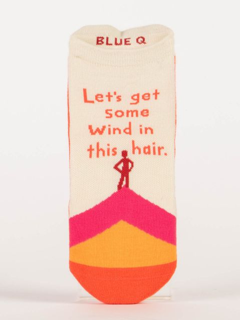 Blue Q sneaker socks. Cream colored with a orange, yellow and pink mountain at the toe of the sock. the silhouette of a little red person stands on the top, hands on hips. Above them, orange text reads "Let's get some wind in this hair" 