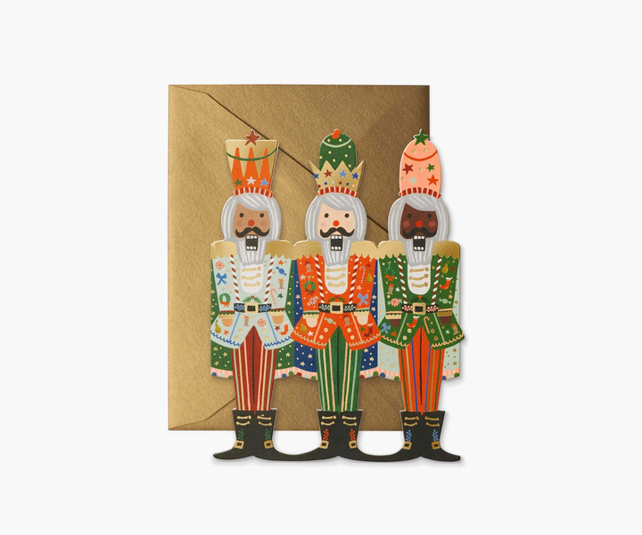 Rifle Paper Co. die cut nutcracker card. Three nutcrackers stand side by side on the front. The first has a light blue jacket and red pants, the second a red jacket and green pants and the last a green jacket with red pants. All of the nutcracker's jackets have small illustrations of other Christmas items. 