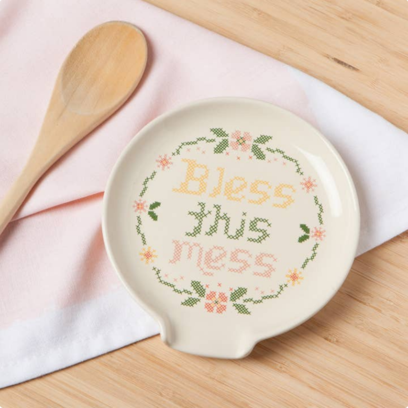 Decorative ceramic dish with cross-stitched text 'Bless this mess' on a wooden surface with a wooden spoon.