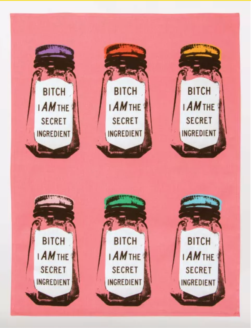 An unfolded Blue Q dish towel. A hot pink towel with 6 black ink, screen printed glass spice jars in two rows of 3. Each has a different colored lid, purple, orange, yellow, beige, green and blue. Each jar has a white label on the front which reads "Bitch I am the secret ingredient"