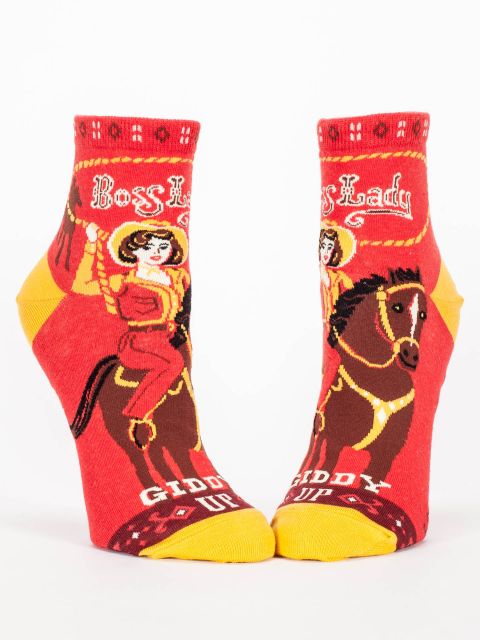 Blue Q Ankle socks. Red with with yellow accents in the heel and toe. The top of the foot has a large illustration of a cowgirl on a brown horse. She has a rope lasso in the air. Inside the lasso in western writing reads "Boss Lady".