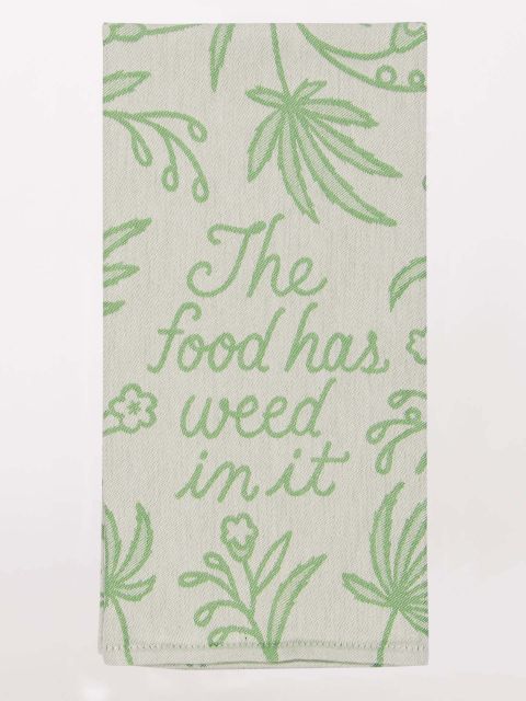 Blue Q dish towel. Light green with a darker green woven botanical pattern containing marijuana leaves. In the middle a script font reads,  "The food has weed in it".