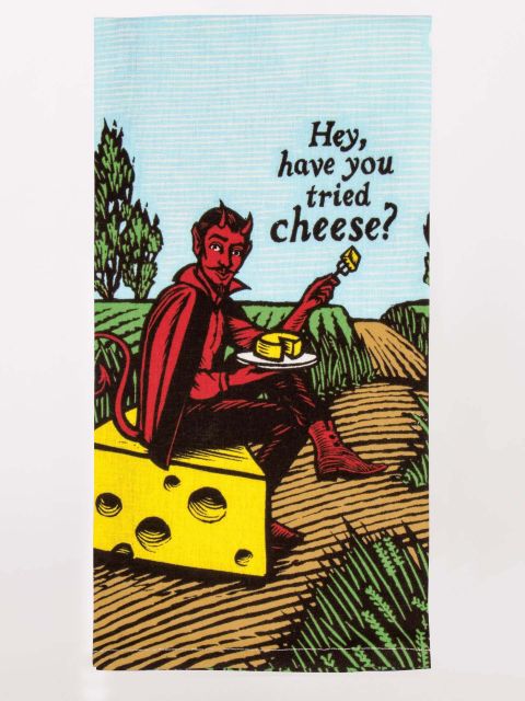 Blue Q screen printed dish towel. an illustrated devil sits on a yellow block of cheese in front of a blue sky and green rolling hills. He's holding a plate of cheese with the words "Hey, have you tried cheese?" above.
