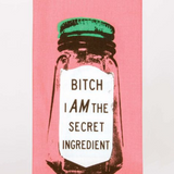 A funny Blue Q dish towel. A hot pink background with a black ink, screen printed image of a glass spice jar. It has a green lid and a white label. The label reads "Bitch I am the secret ingredient".