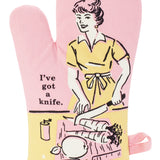 Blue Q oven mitt. Pink, yellow and black illustration of a women cutting up carrots and an apple. Next to her text reads "I've got a knife"