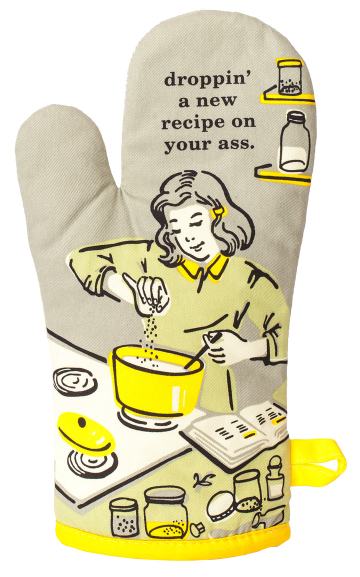 Blue Q oven mitt. A beige background with a black and yellow illustraiton of a girl cooking something in a yellow pot on the stove. At the top it says "droppin' a new recipe on your ass"