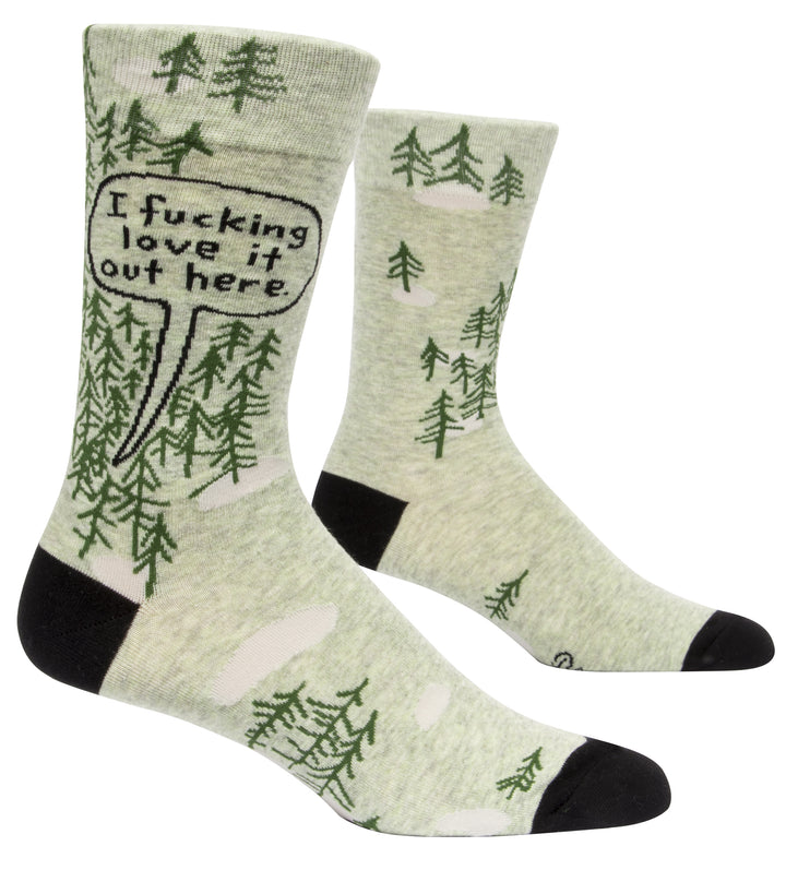 Blue Q crew socks. Light green with black heel and toe. Simple dark green pine tree illustrations dot the upper portion of the sock, a few scattered on the bottom portion of the sock. In a black outlines speech bubble on the top of the sock are the words "I fucking love it out here."